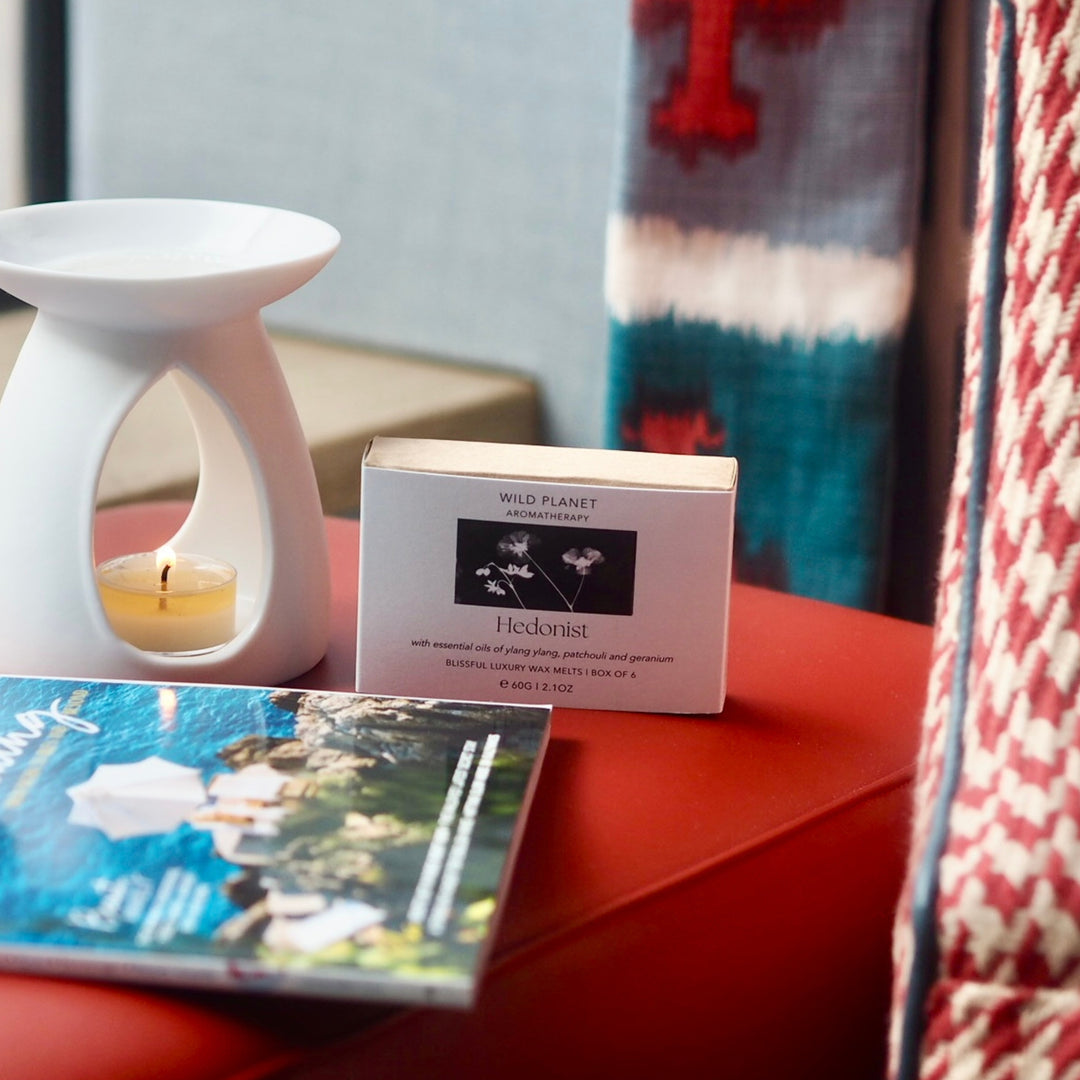 Hedonist Luxury Wax Melt, white ceramic oil burner, lit tea light, box next to magazine on red table