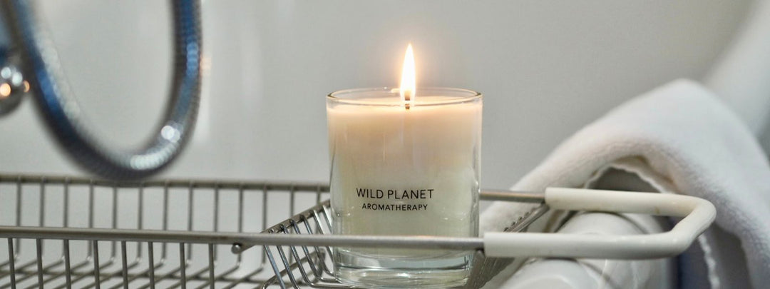 Lit candle on bathroom caddy over bath next to white towel by Wild Planet Aromatherapy