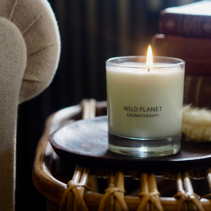 A lit Wild Planet Aromatherapy candle on a wicker table next to a beige chair and a stack of books