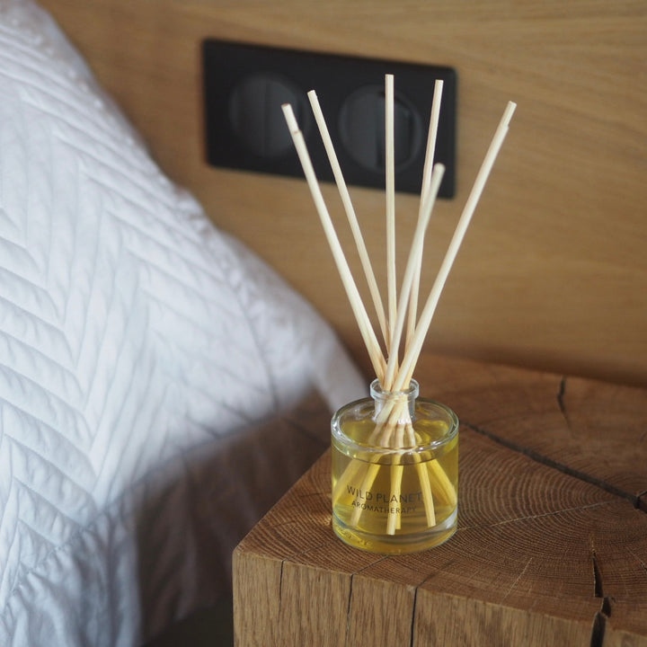 Wild Planet Aromatherapy natural reed diffuser on rustic wooden bedside table near white bedding.