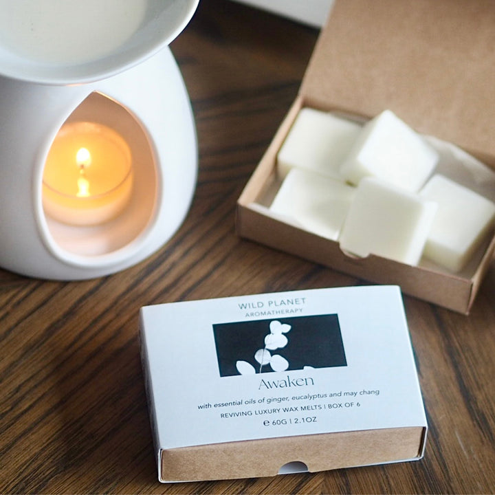 White ceramic oil burner with lit tea light next to box of Wild Planet Aromatherapy luxury wax melts.