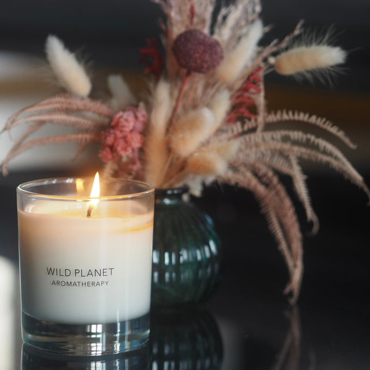 Wild Planet Aromatherapy candle lit on a reflective surface, next to a vase of dried flowers