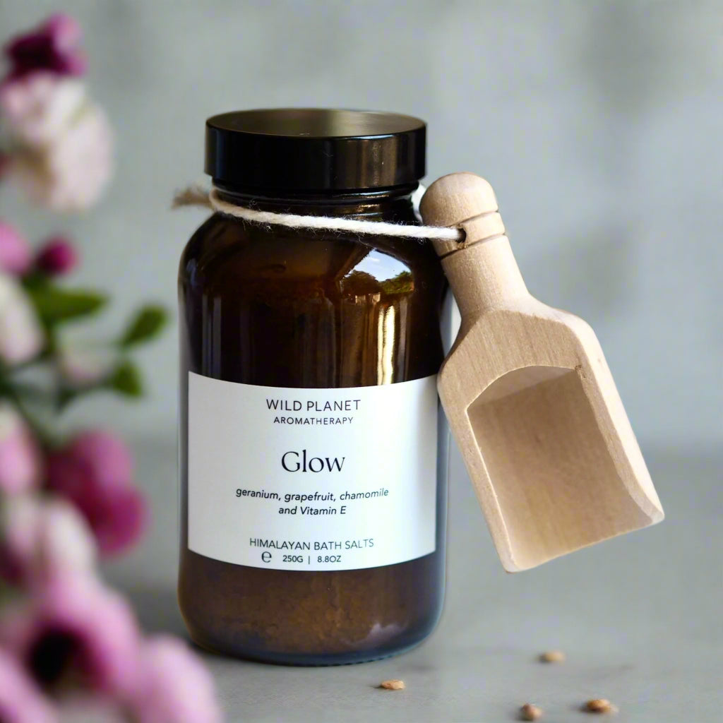 Glow Himalayan bath salts in amber glass jar with wooden scoop on grey background with pink flowers